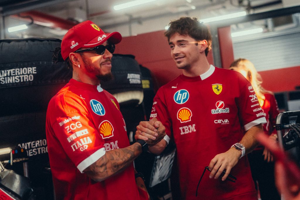 Lewis Hamilton and Charles Leclerc smiling and shaking hands in Ferrari team kit in the garage.